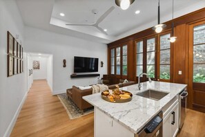 Una cocina moderna con encimera de mármol, horno empotrado y pileta. Hay un comedor con sofá marrón y televisor de pantalla plana en la pared.