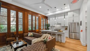 Ein geräumiger Wohnbereich mit einem braunen Sofa, einem Couchtisch und einem raumhohen Fenster mit Holzrahmen. Im Hintergrund ist eine Küche mit weißen Schränken und Edelstahlgeräten zu sehen.