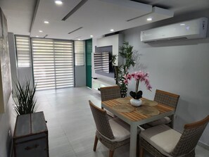 Un espace repas moderne avec une table en bois et des chaises en osier, une plante en pot et une grande fenêtre avec des stores.