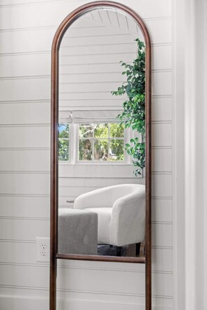 A mirror with a wooden frame reflecting a room with a window, a plant, and a white chair.