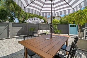 Eine Terrasse mit Holztisch und -stühlen, einem gestreiften Sonnenschirm und einem grauen Zaun.