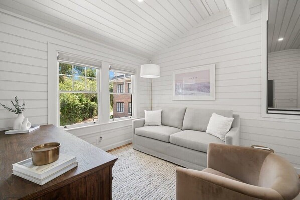 A room with a wooden ceiling, a grey sofa, a brown chair, a white lamp, and a framed picture on the wall.
