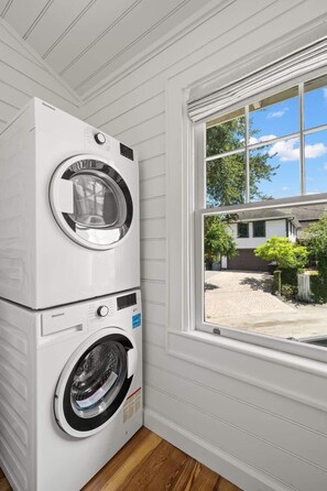 Eine übereinander angeordnete Waschmaschine und ein Trockner in einem Raum mit weißen Wandverkleidungen und einem Fenster mit Blick auf eine Einfahrt.