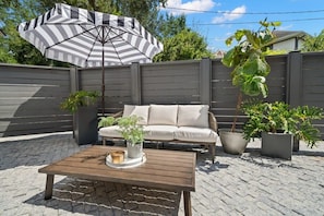 A patio area with a wooden table, a striped umbrella, a beige sofa, and potted plants.