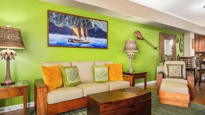 A living room with a green wall, a sofa with orange and green cushions, a wooden coffee table, and a painting of a sailboat. - Hilton Waikoloa pass for 2026. F2 Waikoloa Beach Villas (Waikoloa Beach Resort)