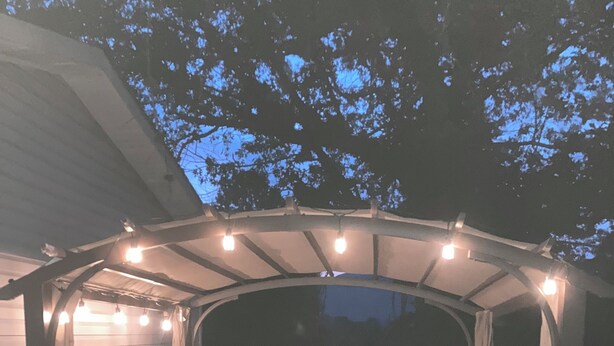 Un patio al aire libre con un brasero, rodeado de sillas y una pérgola con luces colgantes.