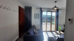 Un salon avec un canapé, une petite table sur laquelle est posée une plante, un ventilateur de plafond et une vue sur un balcon, visible à travers une porte vitrée.
