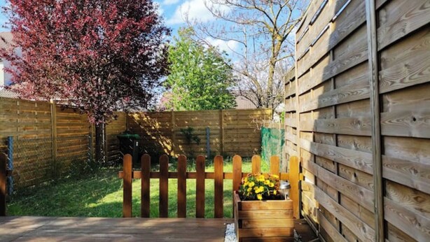 Une terrasse en bois avec une clôture en bois, un arbre aux fleurs roses et un jardinier avec des fleurs jaunes.
