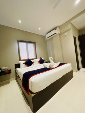 A hotel room with a large bed, a window, an air conditioning unit, and a bedside table with a phone and a red object.