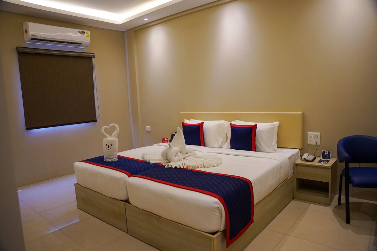 A hotel room with a double bed, a bedside table with a phone and a lamp, a chair, and an air conditioning unit on the wall.