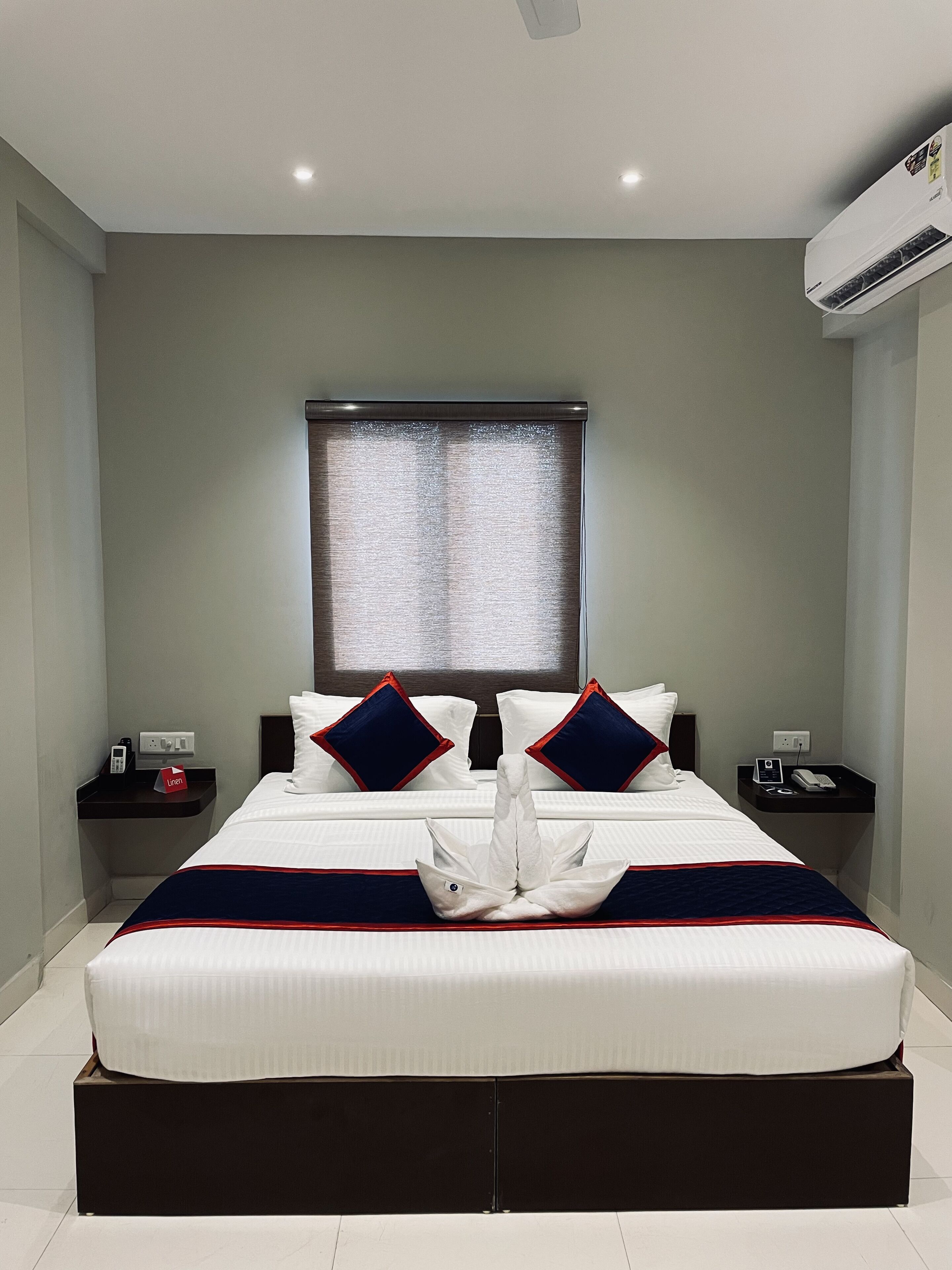A modern hotel room with a large bed, two nightstands, a window with blinds, and an air conditioning unit.