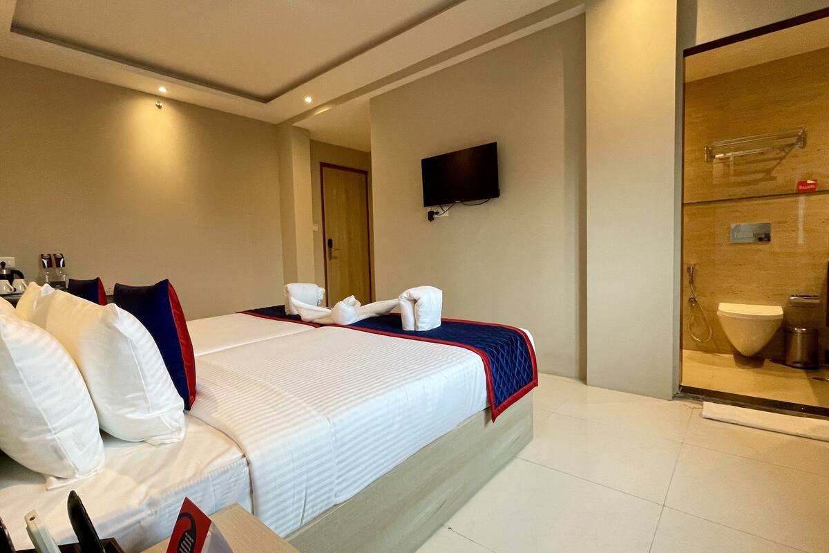 A hotel room with a large bed, a flat-screen TV mounted on the wall, a shower area with a glass door, and a small table with a bottle and glasses.