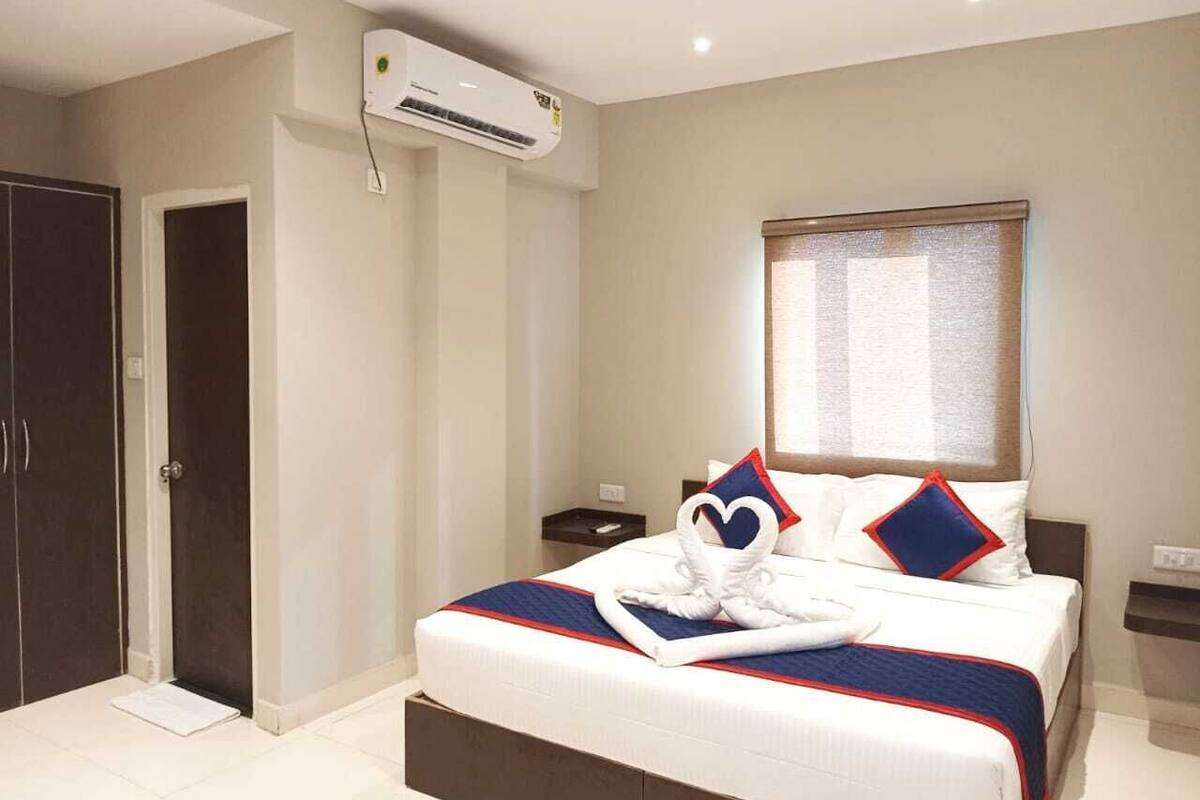 A hotel room with a large bed, a wardrobe, an air conditioning unit, a bedside table, and a window with blinds.