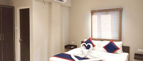 A hotel room with a large bed, a wardrobe, an air conditioning unit, a bedside table, and a window with blinds.