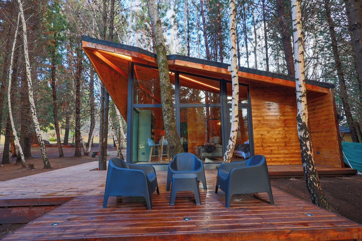 Une cabane moderne en bois, dotée de grandes fenêtres et d’une terrasse avec des chaises.