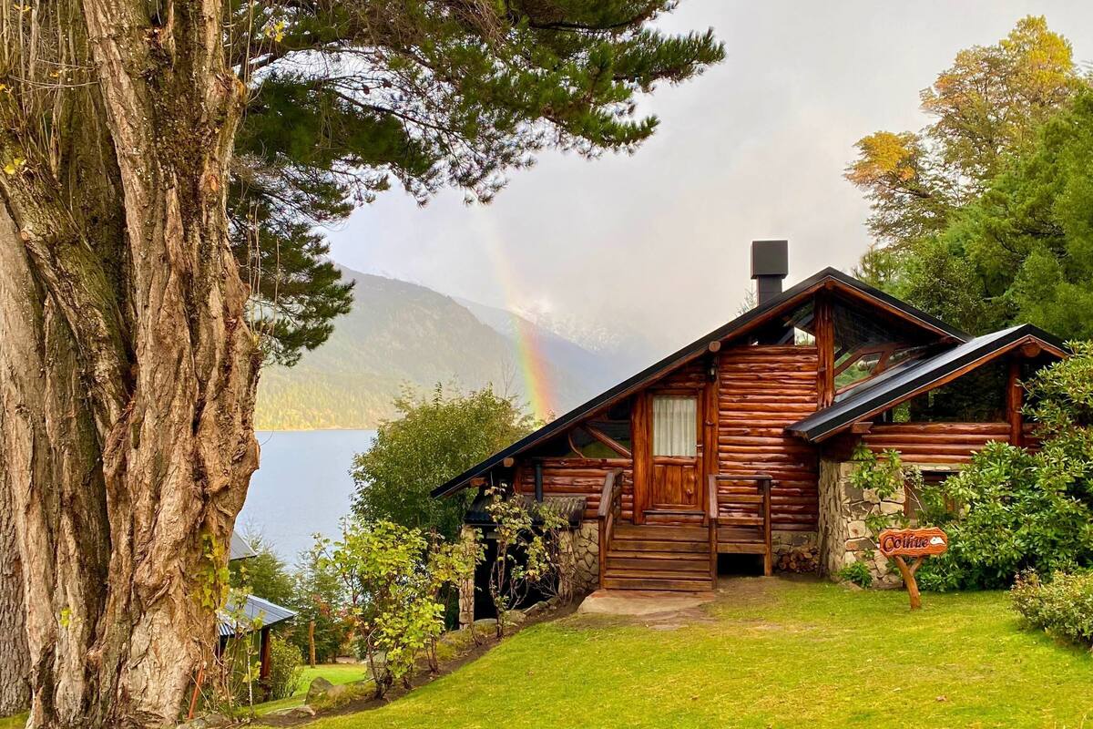 Une cabane rustique en rondins de bois, avec des fondations en pierre, une véranda en bois et une enseigne indiquant le nom « Colline ».