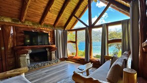 A rustic cabin interior with a stone fireplace, wooden furniture, and a large window offering a view of a lake. - Villa Labrador Bariloche (San Carlos de Bariloche)