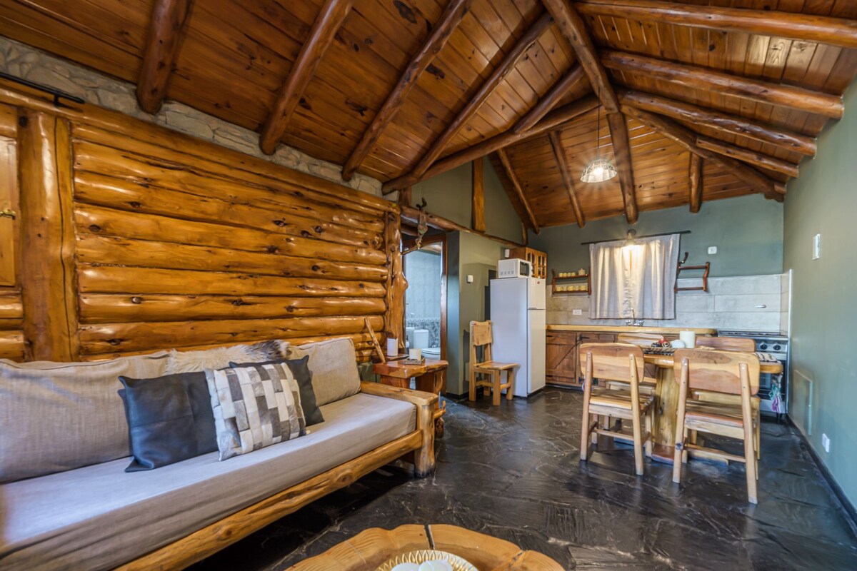 Een inrichting van een blokhut met houten plafond, een bank met kussens, een eettafel met stoelen, een keuken met een koelkast en magnetron, en een stenen open haard.