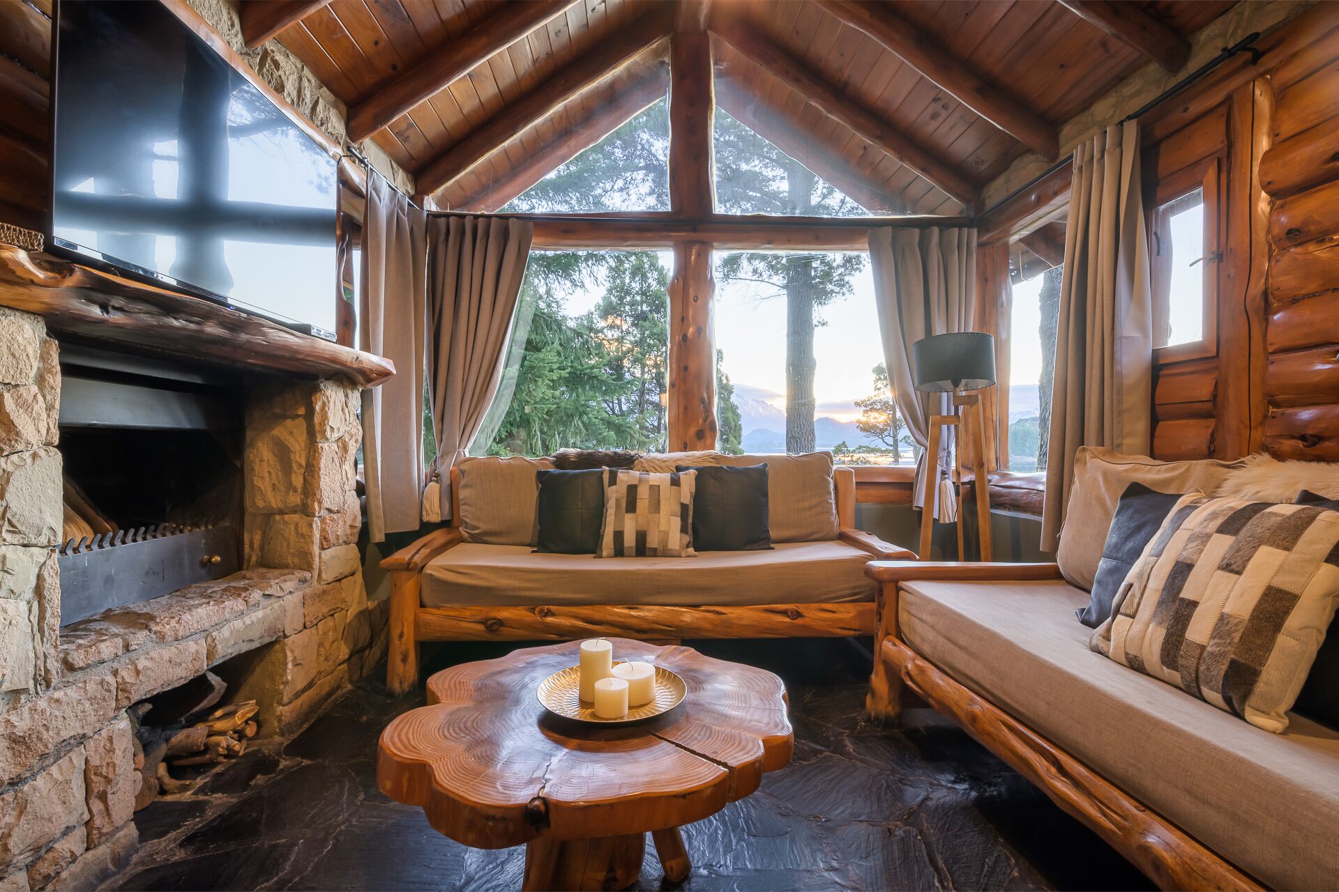 Interior acogedor de una cabaña con chimenea de piedra, muebles de madera y ventanas amplias con vistas a árboles y montañas.