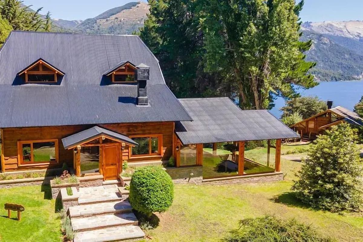 Une grande cabane en rondins avec un toit en pente, deux lucarnes et un porche couvert.
