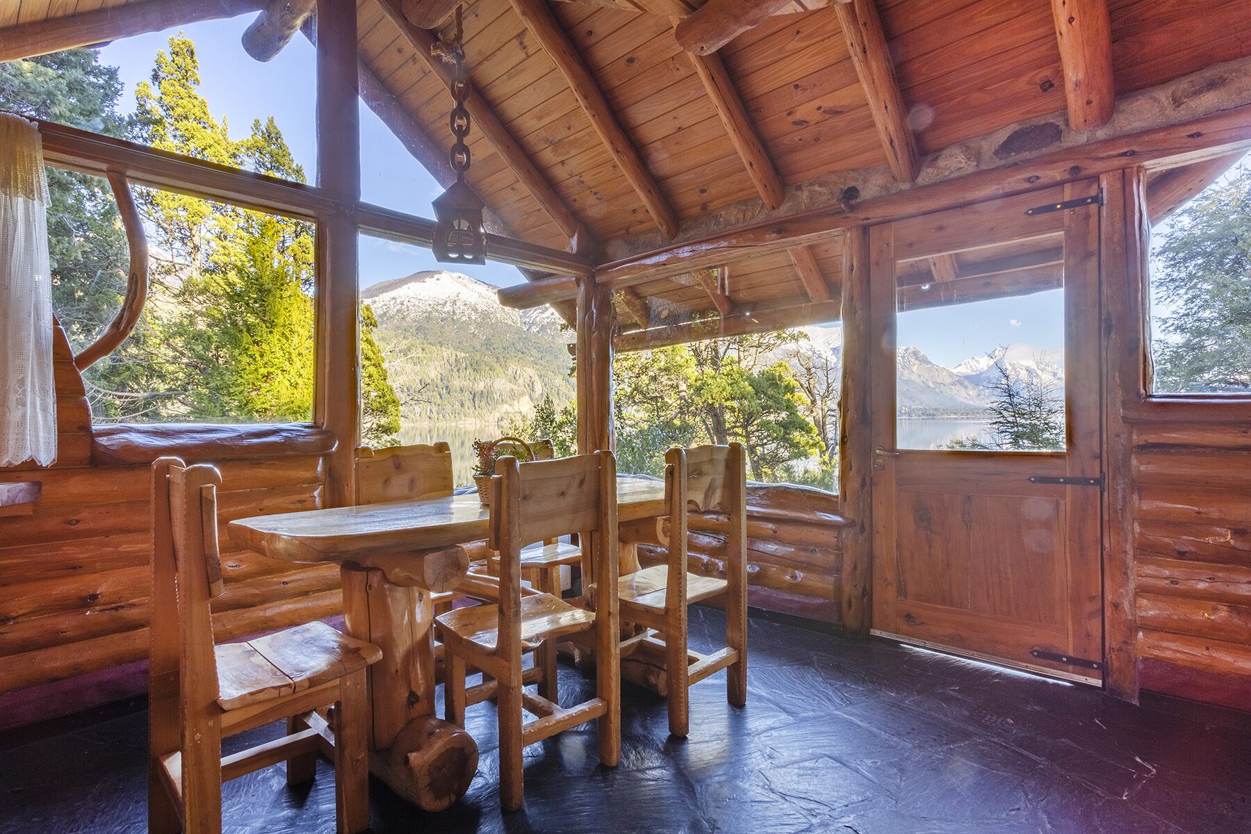 A wooden cabin with a dining area featuring a table and chairs, large windows offering mountain views, and a rustic interior design.