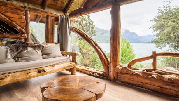 A wooden cabin with a sofa and a round wooden table, large windows with a view of a lake and mountains. - Villa Labrador Bariloche (San Carlos de Bariloche)