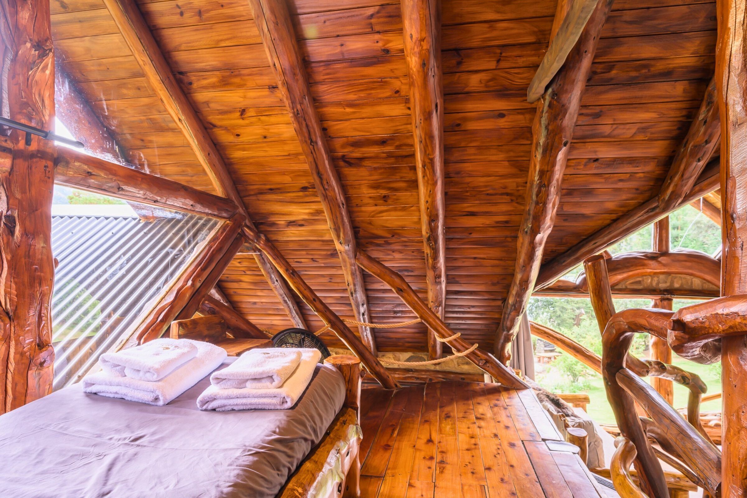 Una cabaña de madera con techo inclinado, una cama con toallas dobladas y vistas al verdor exterior.