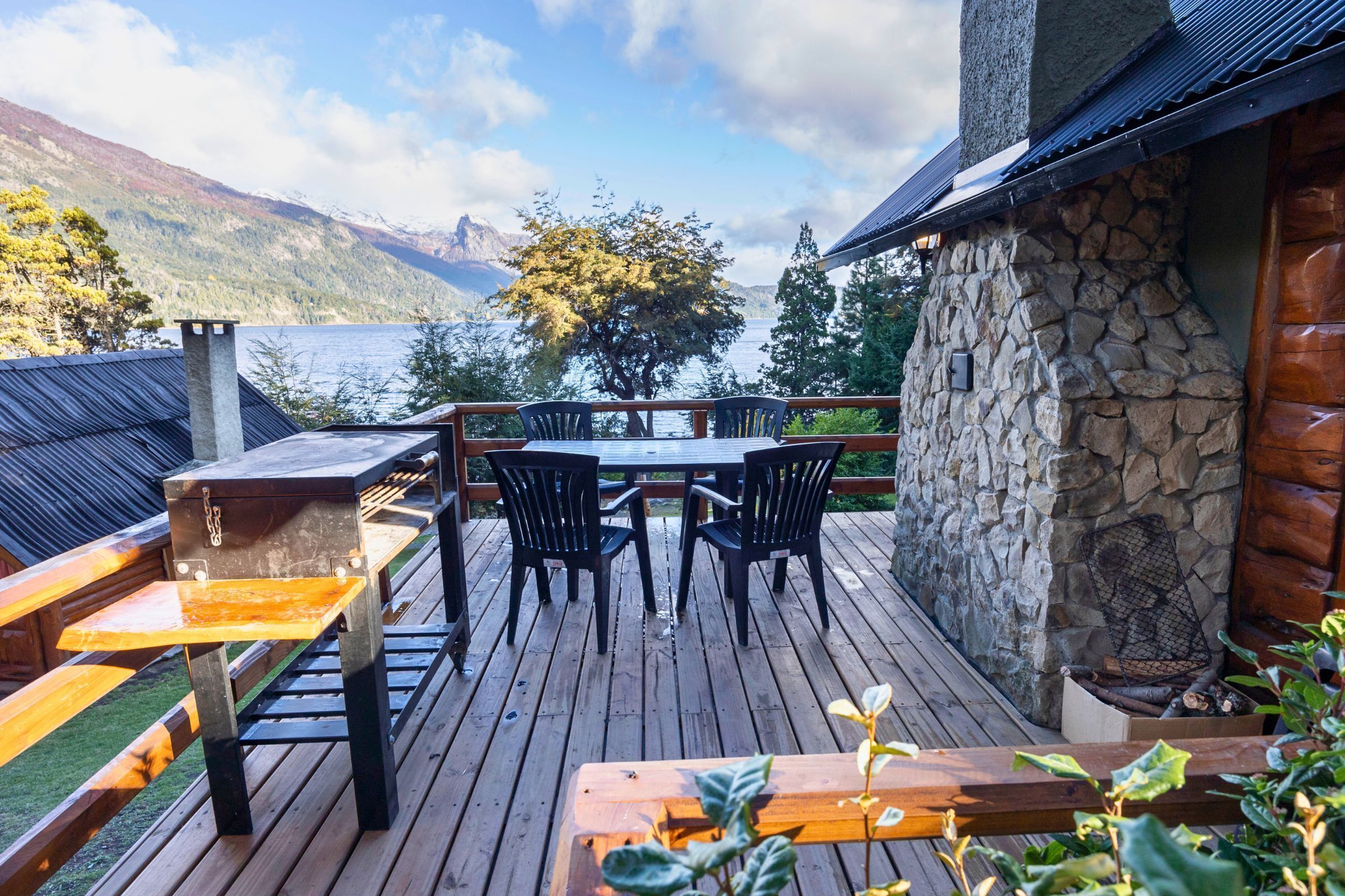 A wooden deck with a stone wall, a table with chairs, and a view of a lake and mountains.