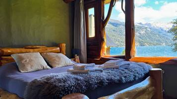 A wooden cabin bedroom with a bed, pillows, a fur throw, and a view of a lake and mountains.