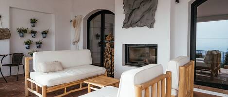 A modern living room with a white sofa, wooden chairs, a small table, and a fireplace. There are potted plants on the wall and a view through an arched doorway.