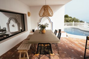 A dining area with a table, chairs, and a bench. There is a large window with a decorative mirror and a view of a swimming pool.