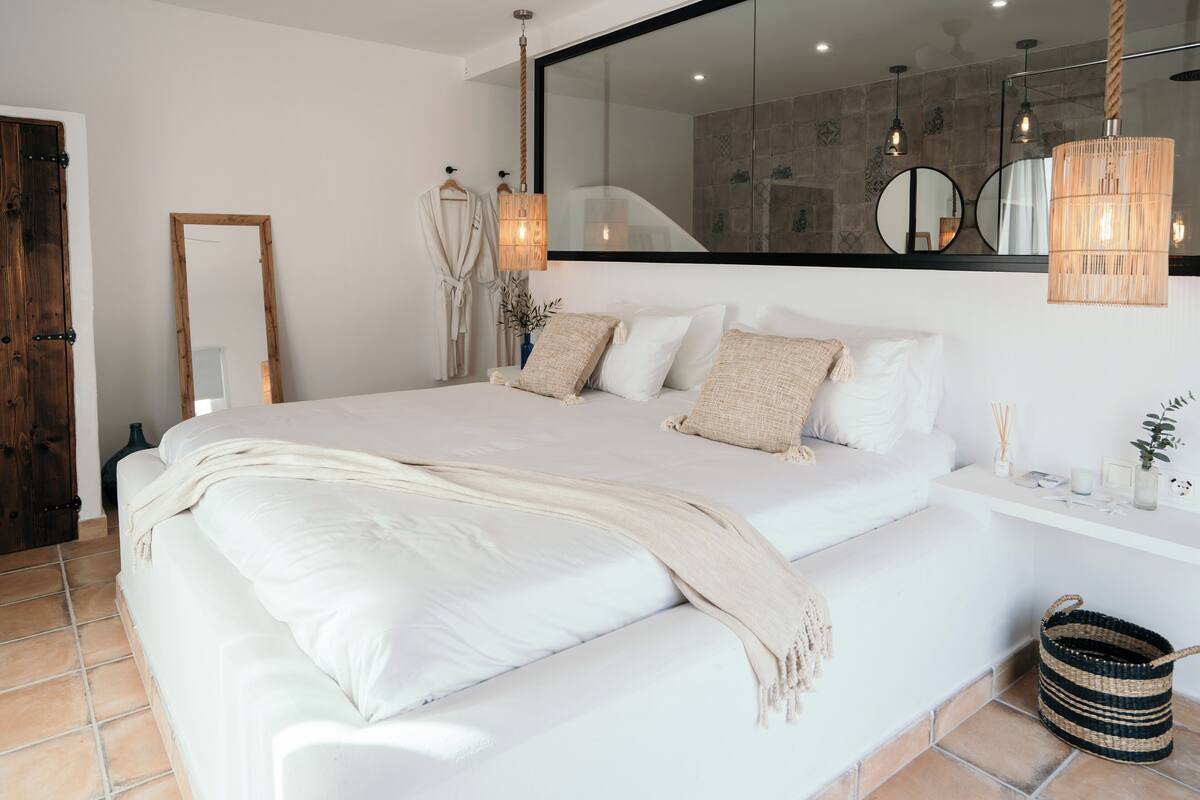 A modern bedroom with a large bed, a wooden door, a mirror, and a hanging light fixture.