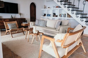 Ein Wohnzimmer mit einer Couch, zwei Holzstühlen, einem weißen Teppich, einem Holztisch, einem Bücherregal und einer Treppe.