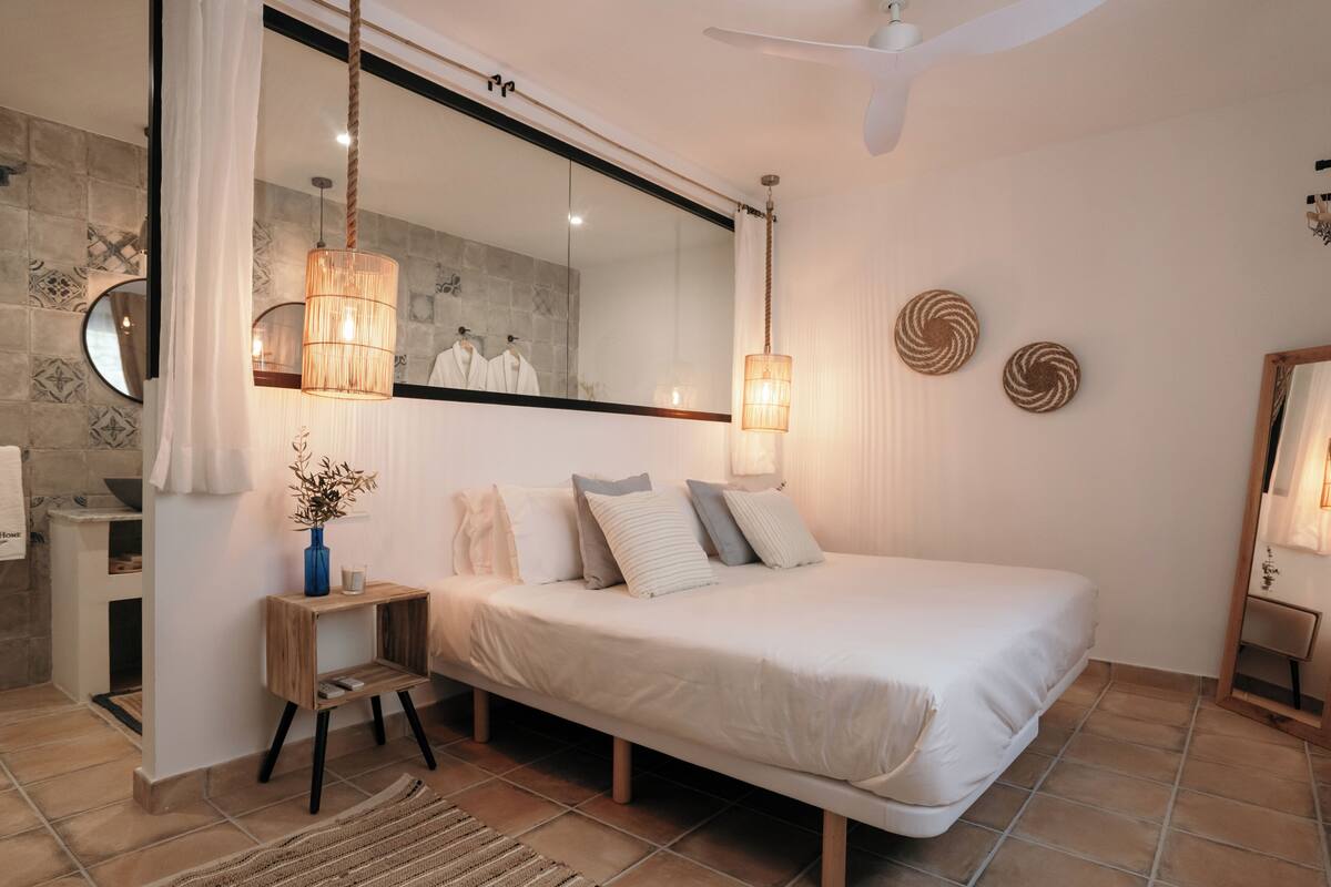 A bedroom with a large bed, a bedside table, a mirror, and a hanging light fixture.