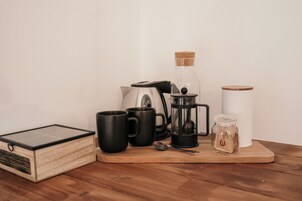 A wooden table with a coffee maker, two black mugs, a spoon, a glass jar, and a white container.