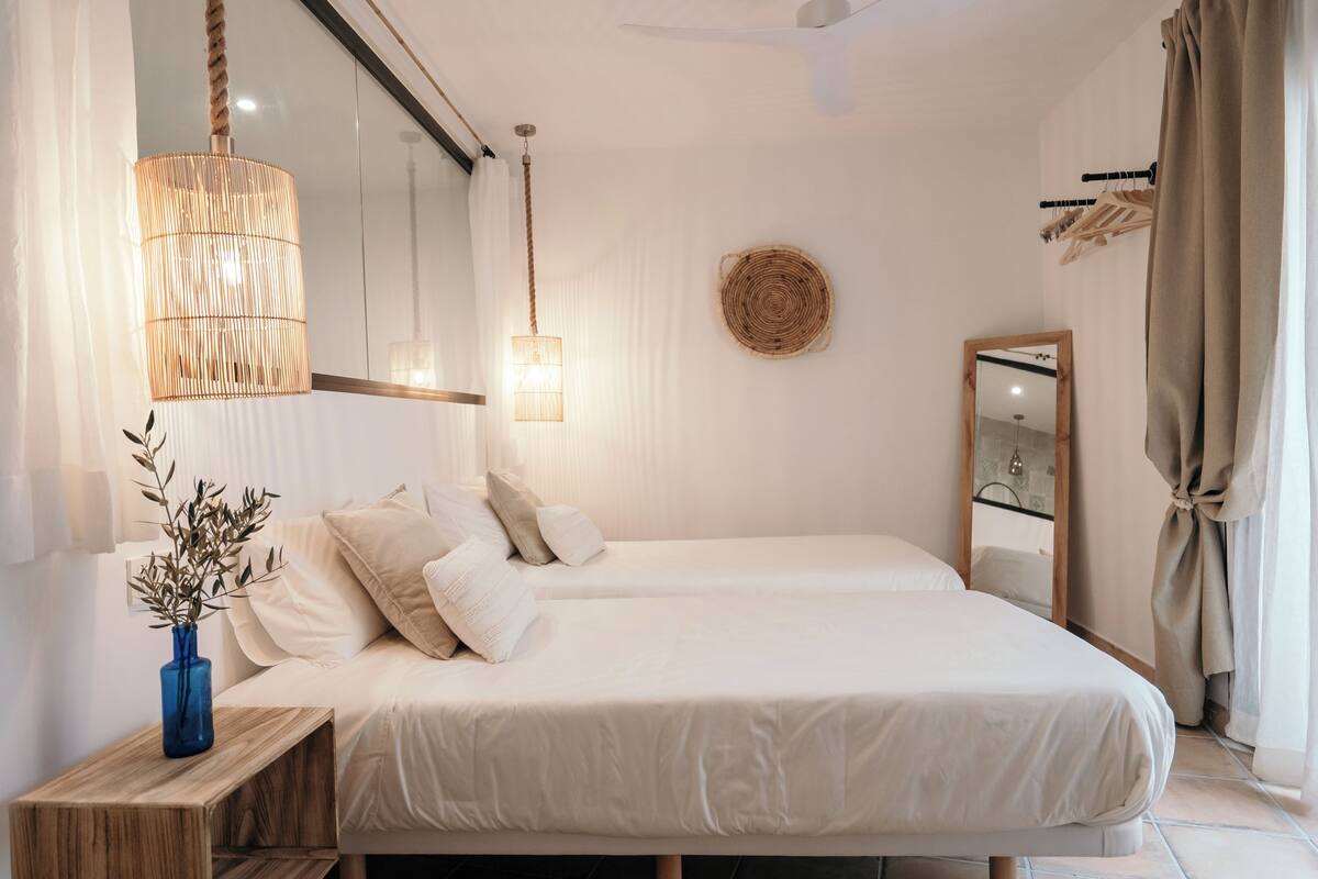 A modern bedroom with two beds, a bedside table with a vase, a hanging light fixture, a mirror, and a woven wall decoration.