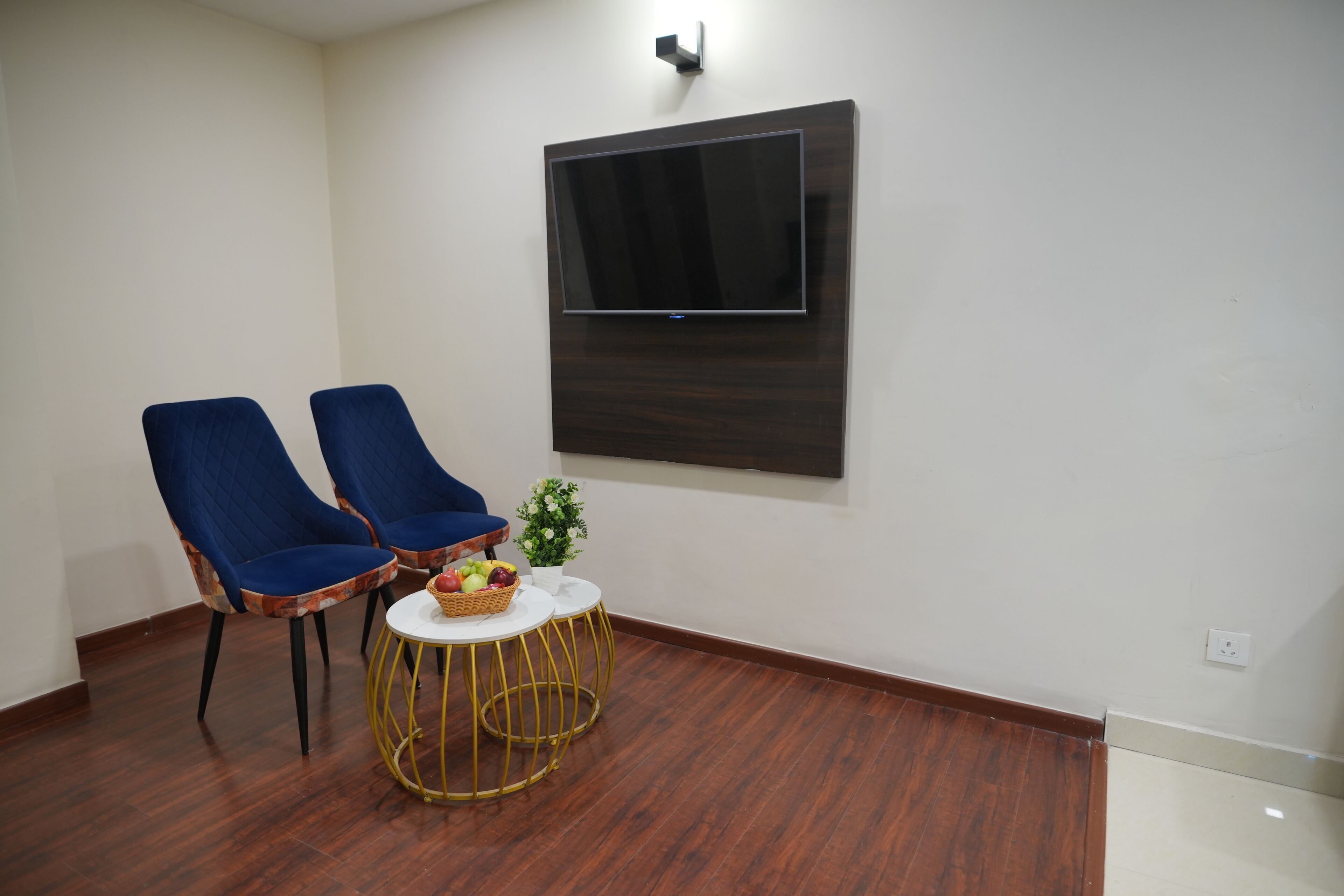 Een kamer met twee blauwe stoelen, een kleine ronde tafel met een mand fruit, en een wandgemonteerde flatscreen televisie.