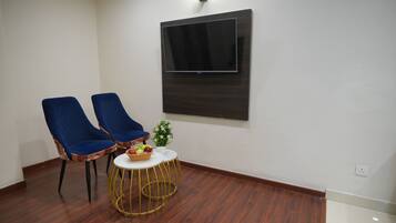 Een kamer met twee blauwe stoelen, een kleine ronde tafel met een mand fruit, en een wandgemonteerde flatscreen televisie.