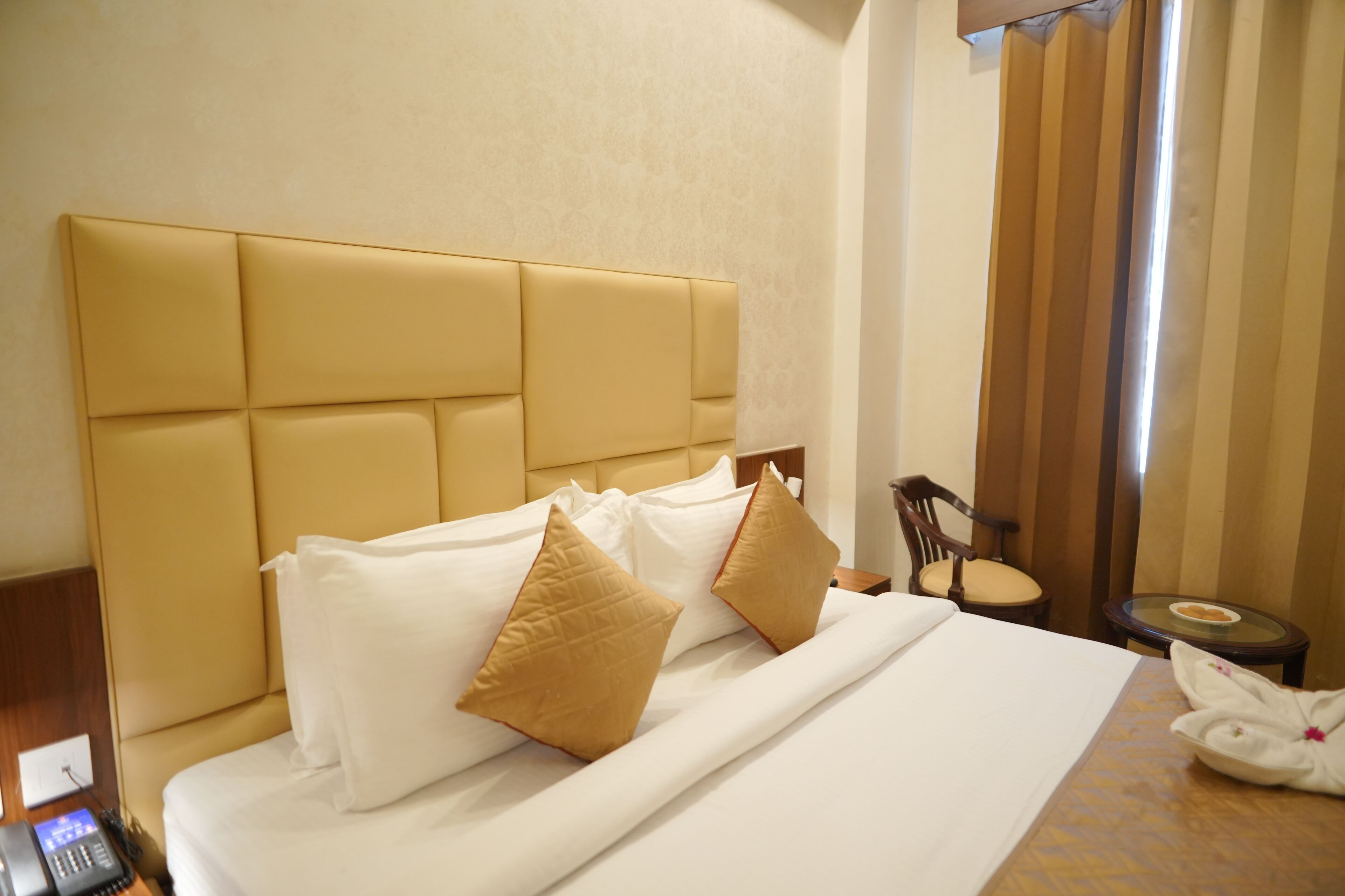 A hotel room with a large bed, two pillows, a bedside table, a chair, and a small table with a glass top.