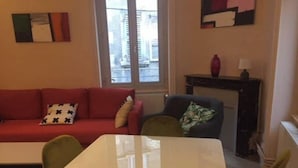 Ett vardagsrum med en röd soffa, gröna fåtöljer, ett vitt bord med en fruktkorg och ett fönster med utsikt över byggnader.