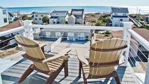 Een balkon met twee Adirondack-stoelen en een kleine tafel, met uitzicht op een strand en huizen.