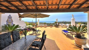 Um pátio externo com mesa e cadeiras, um grande guarda-sol e plantas em vasos.