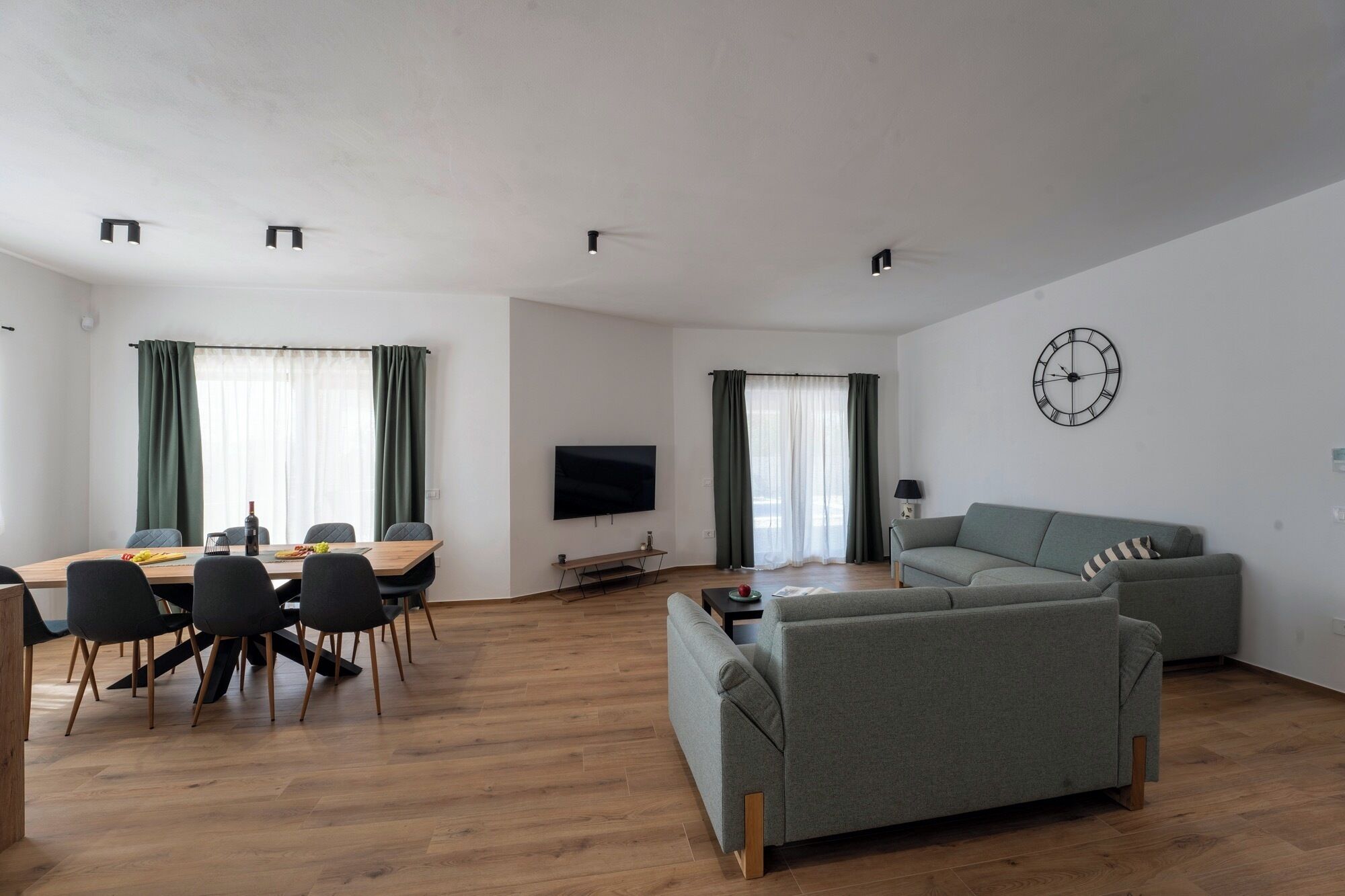 Un soggiorno moderno con un divano grigio, un tavolo da pranzo con sedie, una TV a schermo piatto montata a parete e una grande sveglia.