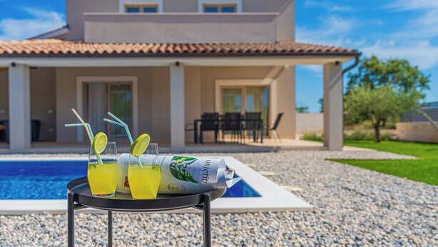 Ein Tisch am Pool mit Getränken und einer Zeitschrift, ein Schwimmbecken und ein Haus mit einem Ziegeldach und zwei Fenstern.
