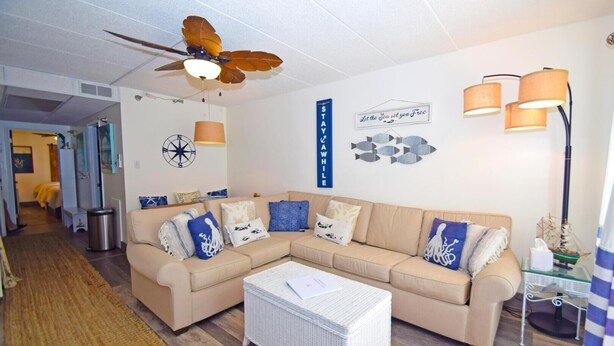 A living room with a beige sectional sofa, a white wicker ottoman, a ceiling fan, wall-mounted lamps, and nautical-themed decor.