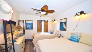 A room with two beds, a ceiling fan, artwork on the walls, and a desk with a lamp and a jar of seashells.