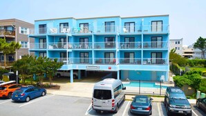 A multi-story hotel with a blue exterior, a pool, and a parking area with various vehicles.