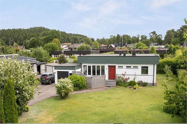 A single-story house with a green roof, a red door, and a driveway leading to a parked car. - Vistelse House (Hallands län)