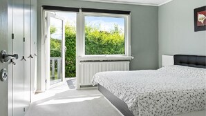 Une chambre avec un lit, un radiateur, une fenêtre donnant sur un espace verdoyant et une porte menant à un espace extérieur.