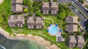 Eine Vogelperspektive zeigt ein Resort mit mehreren bungalowartigen Gebäuden, einem zentralen Swimmingpool und einem Strand in unmittelbarer Nähe.